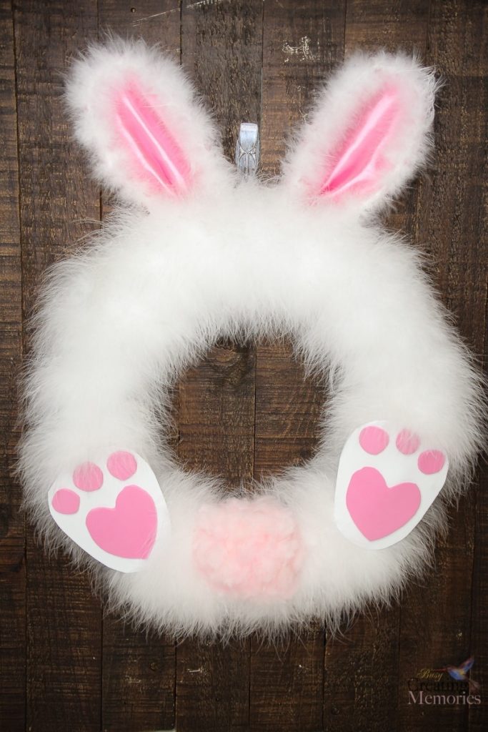 Easter wreath made of white feather boa and with bunny ears, feet, and fluffy tail, on a dark background.